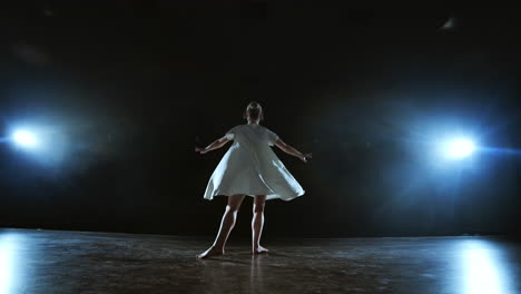 Zoom-camera-moves-around-the-stage-with-software-and-smoke.-Girl-ballerina-dancing-in-a-white-dress-spinning-plastic-while-performing-pirouettes-and-rotations-experiencing-emotions-and-dramatic-atmosphere.